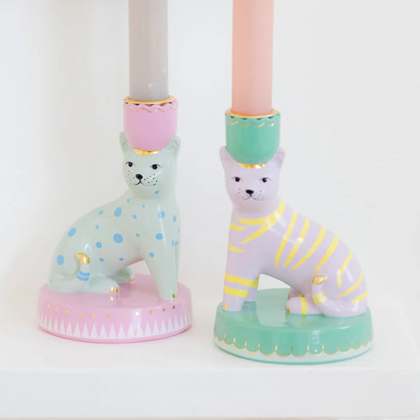 Two candlestick holders with a cat design, one in pink and one in green, with gold detailing, on a white surface.